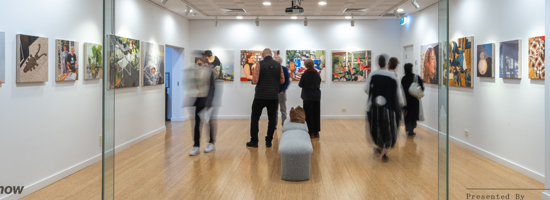Visitors mingling in a well lit gallery space with paintings hung on the wall, observed through a glass window